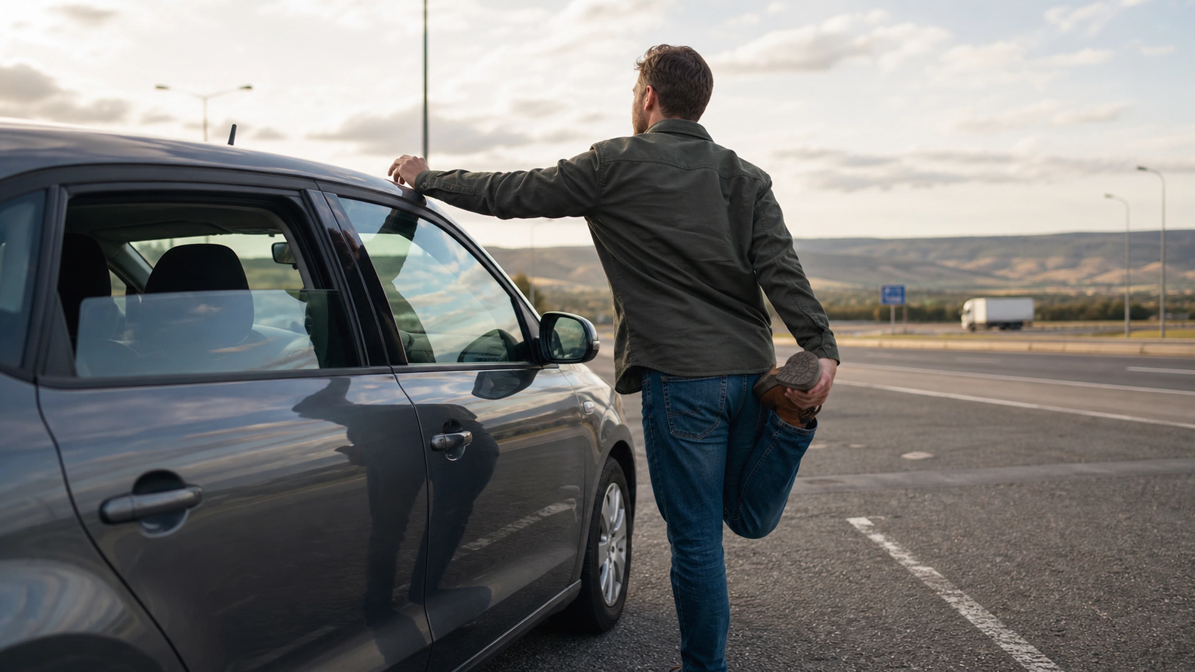 Driver taking a standing hip and lower-back stretch beside a parked car at a motorway rest stop