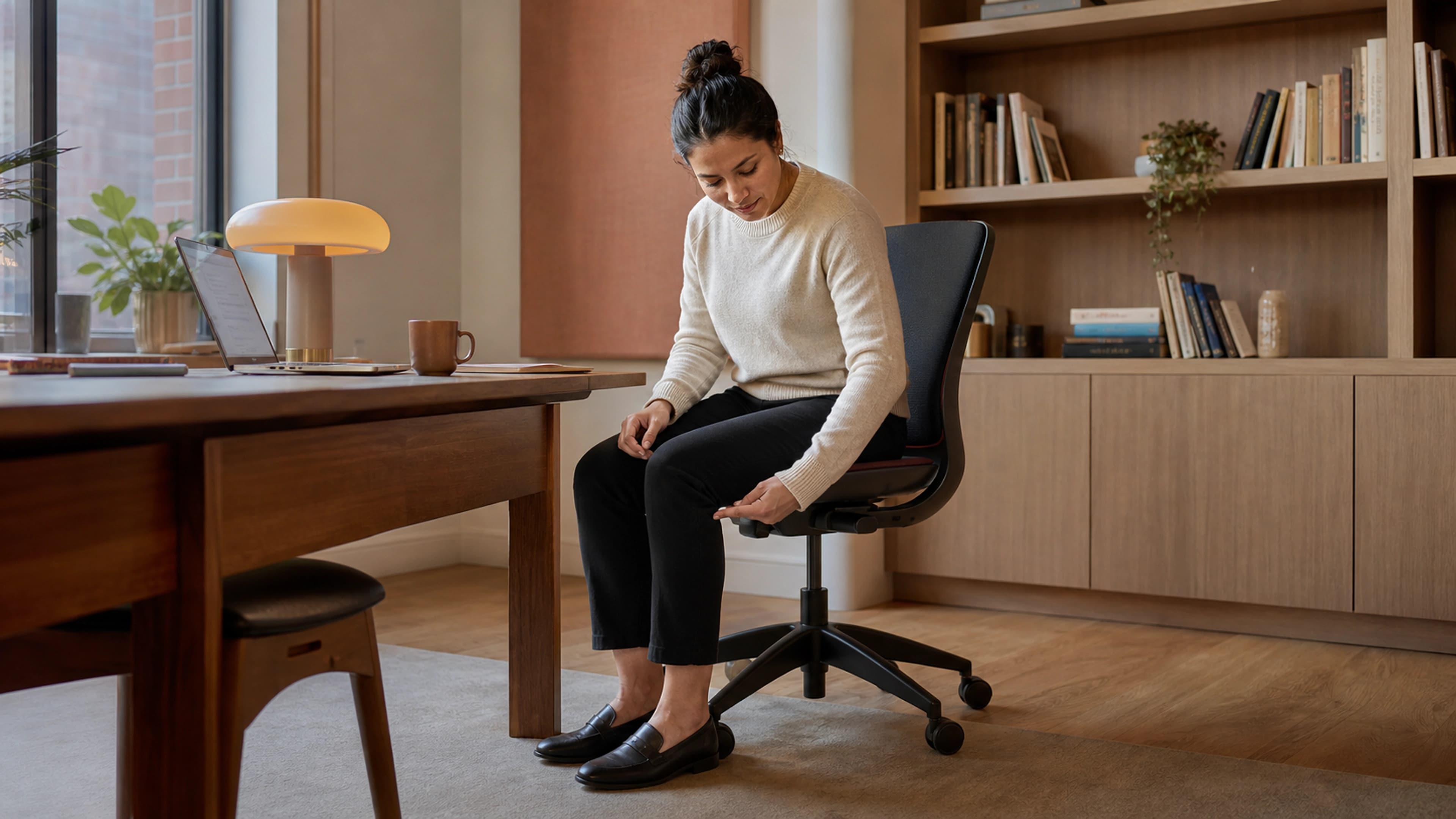 Office worker assessing seated alignment and chair depth from a side angle in a daylight workspace