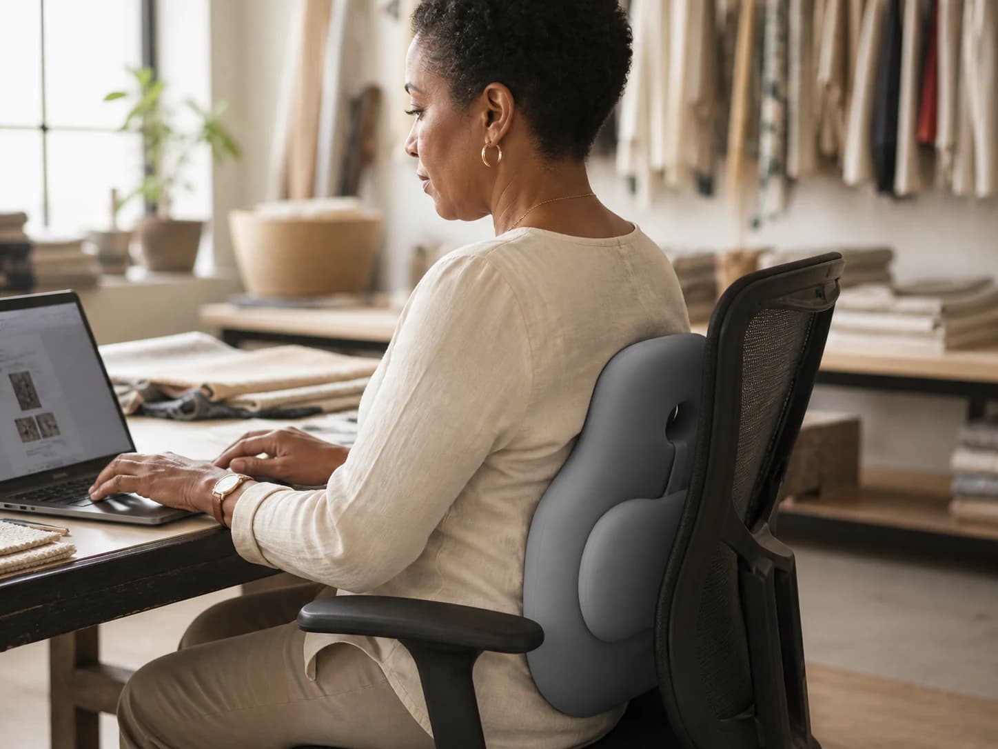 Seated adult using the volcanic-gray HaloBack cushion at a textile-studio worktable